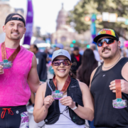 Three smiling runners pose outdoors in the summer sun, each holding a race medal. They wear athletic gear and sunglasses, proudly showing their commit to fitness with a blurred crowd and building in the background. Austin Marathon Half Marathon & 5K