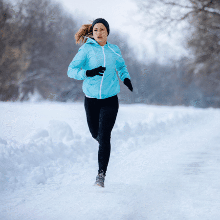 A woman in a light blue jacket, black leggings, and gloves runs on a snowy path in a winter landscape, surrounded by snow-covered trees, demonstrating the importance of hydration strategies even on long runs in cold weather. Austin Marathon Half Marathon & 5K